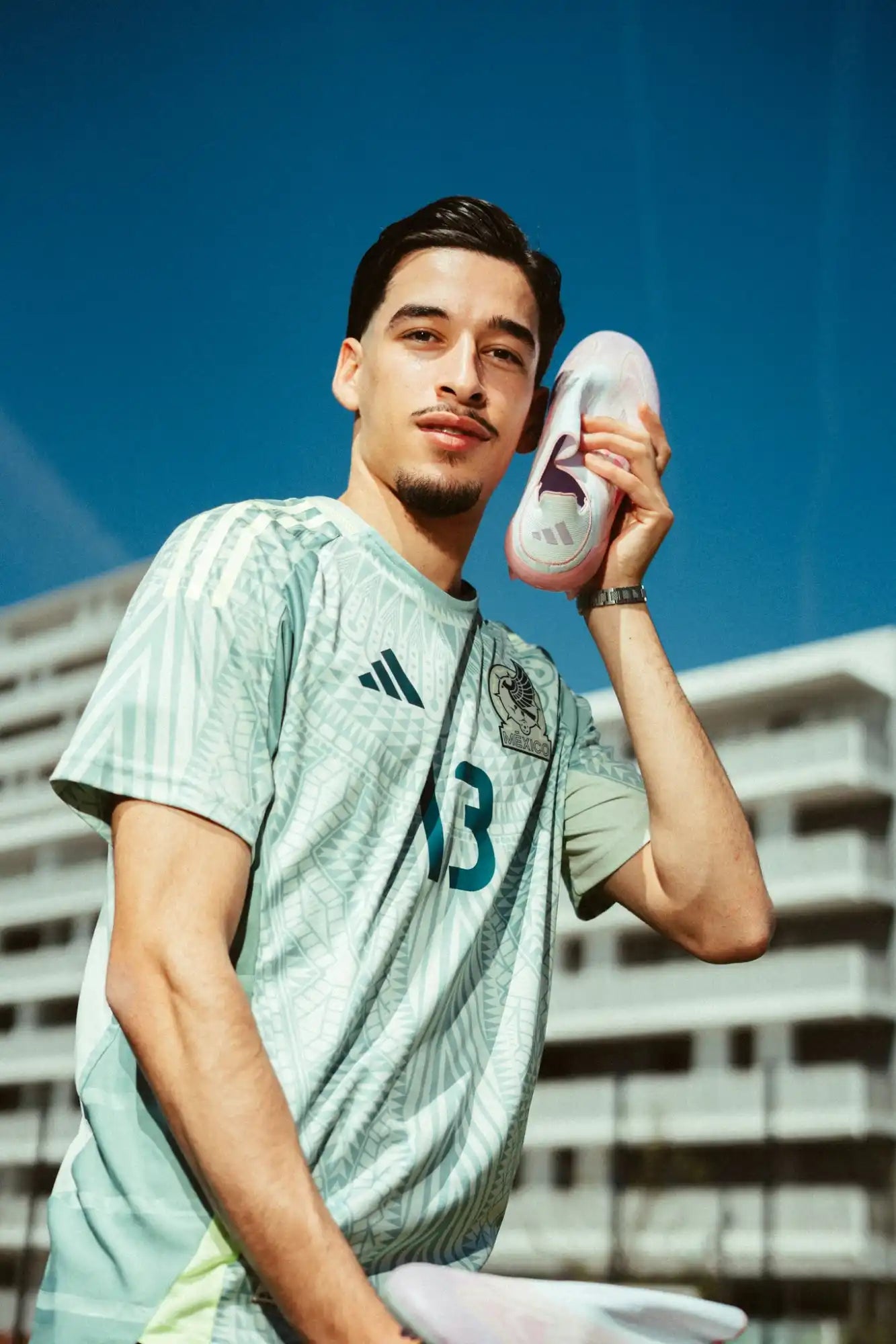 Image of Un homme portant une chemise à manches courtes à motif vert clair et blanc avec un logo adidas noir et le numéro 13, tenant une chaussure de sport rose clair et blanche à l'oreille.