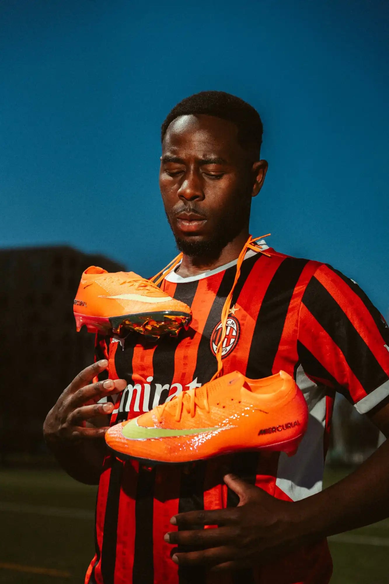 Homme portant un maillot rayé rouge et noir tenant deux crampons de football orange vif.
