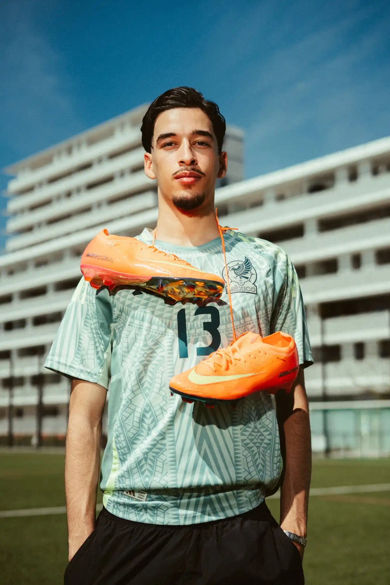 Un jeune homme portant un maillot vert clair avec le numéro 13, tenant une paire de crampons de football orange vif autour du cou.