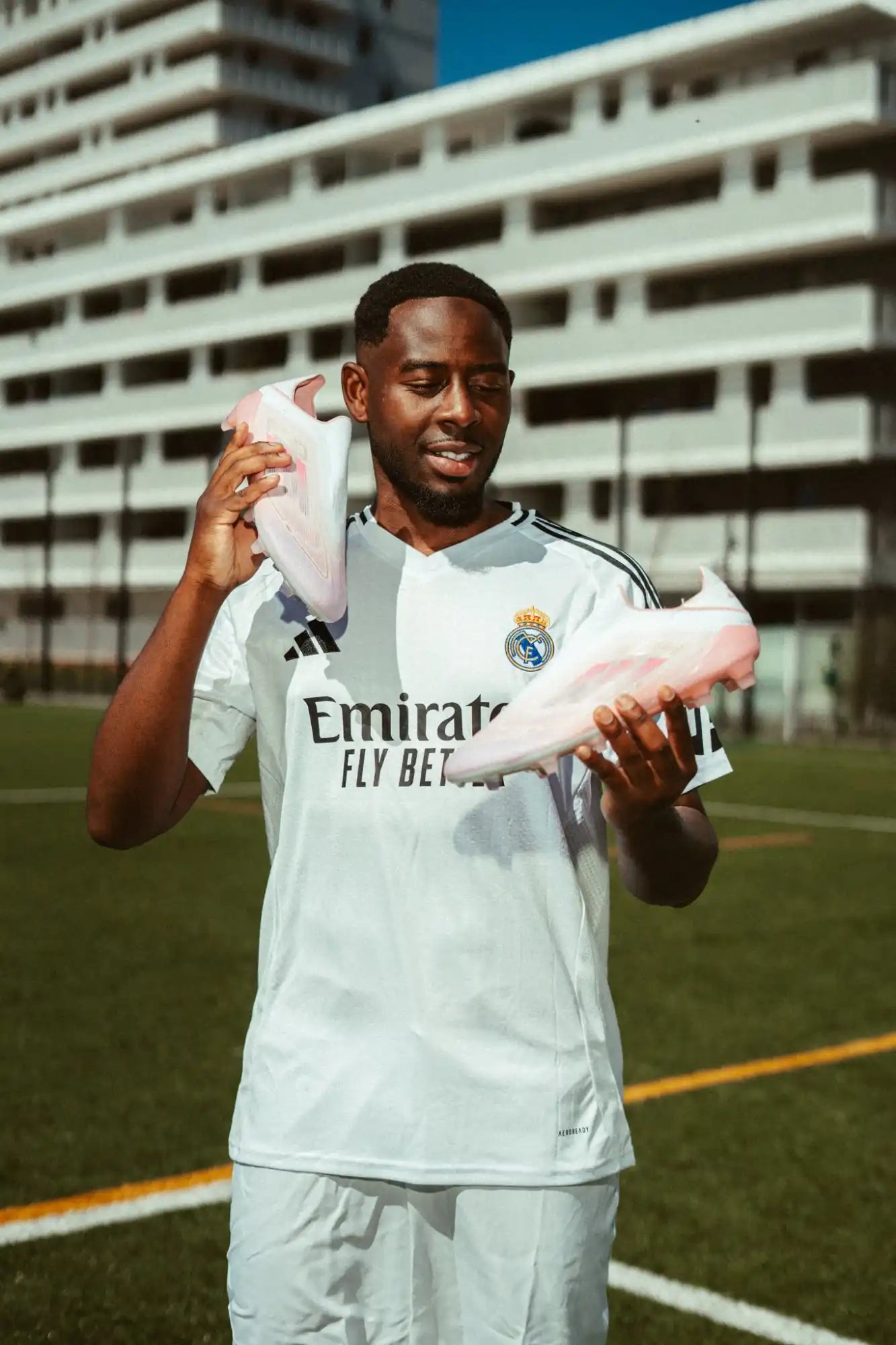 Un joueur de football masculin en maillot blanc du Real Madrid tenant deux paires de chaussures de football rose clair.